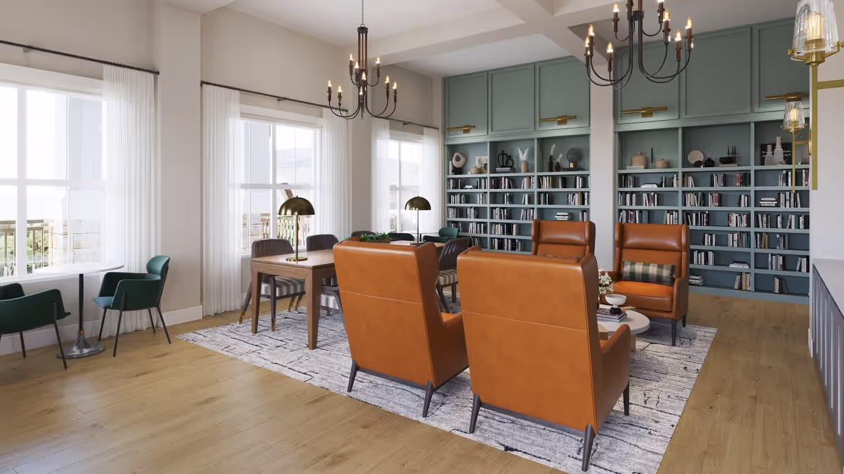 A bright and spacious living room with large windows covered by sheer white curtains. The room features a wooden floor, a large area rug, two brown leather armchairs around a small coffee table, a wooden table with chairs, green armchairs near the windows, and a built-in bookshelf filled with books and decorative items. Two black chandeliers hang from the ceiling.