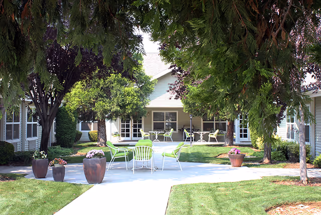Outdoor courtyard area at Avista Senior Living Nampa featuring a concrete patio with green chairs and white tables, surrounded by grass, trees, and potted flowers, with building entrances visible in the background.