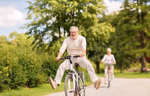 An elderly man rides a bicycle with his legs outstretched along a tree-lined path while an elderly woman cycles behind him.