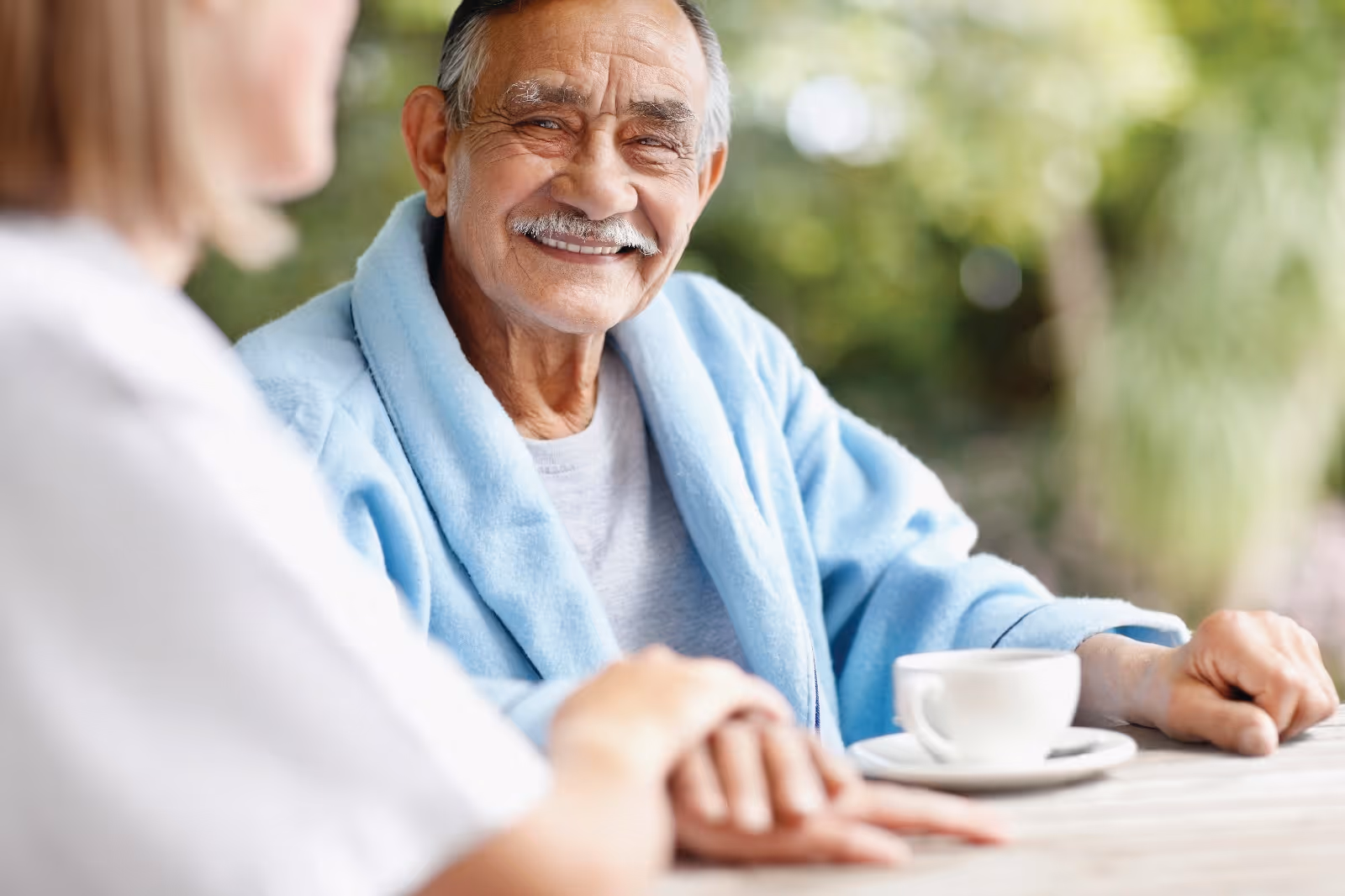 Smiling elderly man in a blue robe sits at an outdoor table with a coffee cup.