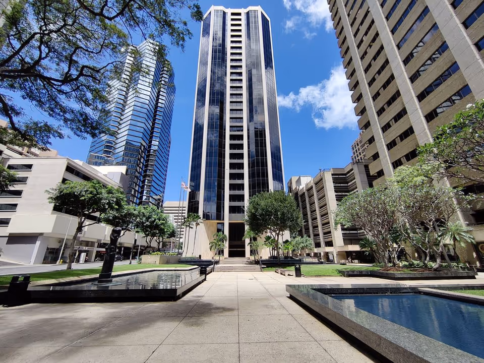 A wide outdoor plaza with tall modern buildings, trees, and reflective water features.