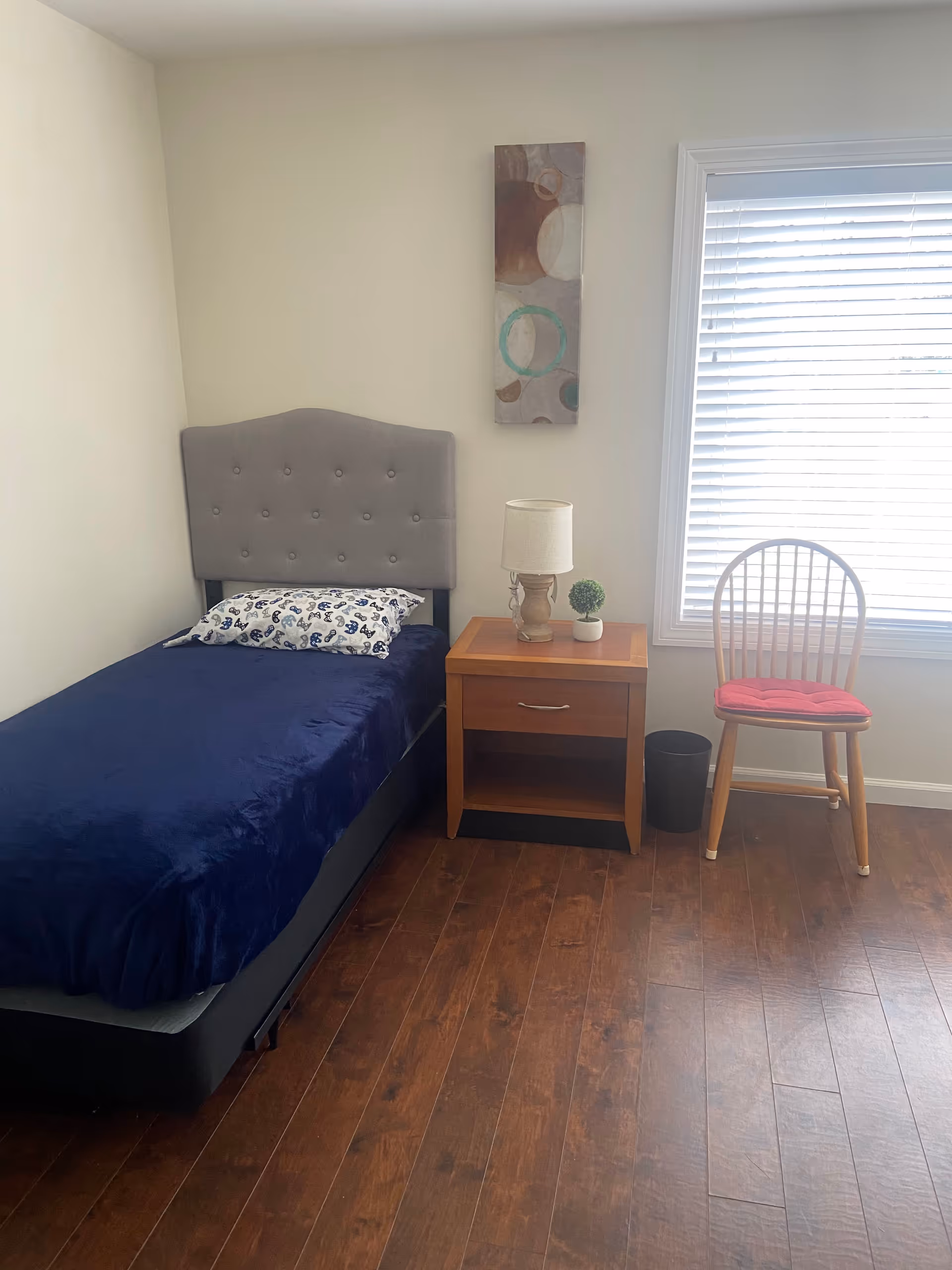 A bedroom with a single bed, blue linens, nightstand, lamp, and a chair by the window.
