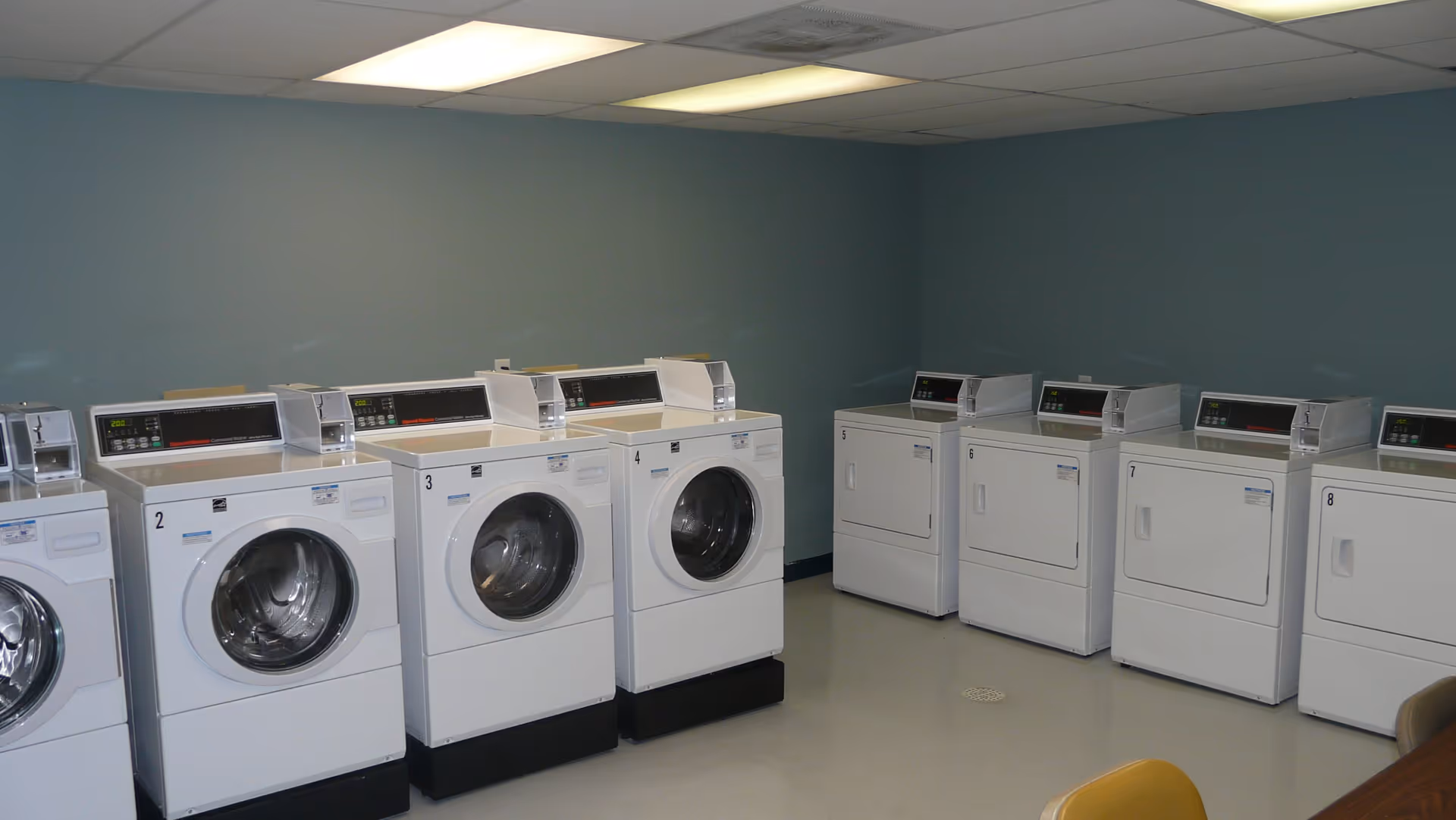 Communal laundry room with front-loading washers and dryers arranged along teal walls under fluorescent ceiling lights.