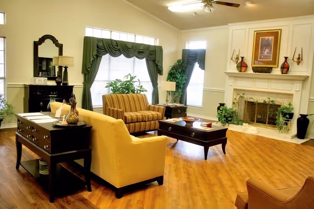 A bright and spacious living room with wooden flooring, featuring a yellow armchair, a striped loveseat, a dark wooden coffee table, and a fireplace with decorative items on the mantel. The room has large windows with green drapes, a ceiling fan, and various plants and lamps adding to the cozy atmosphere.