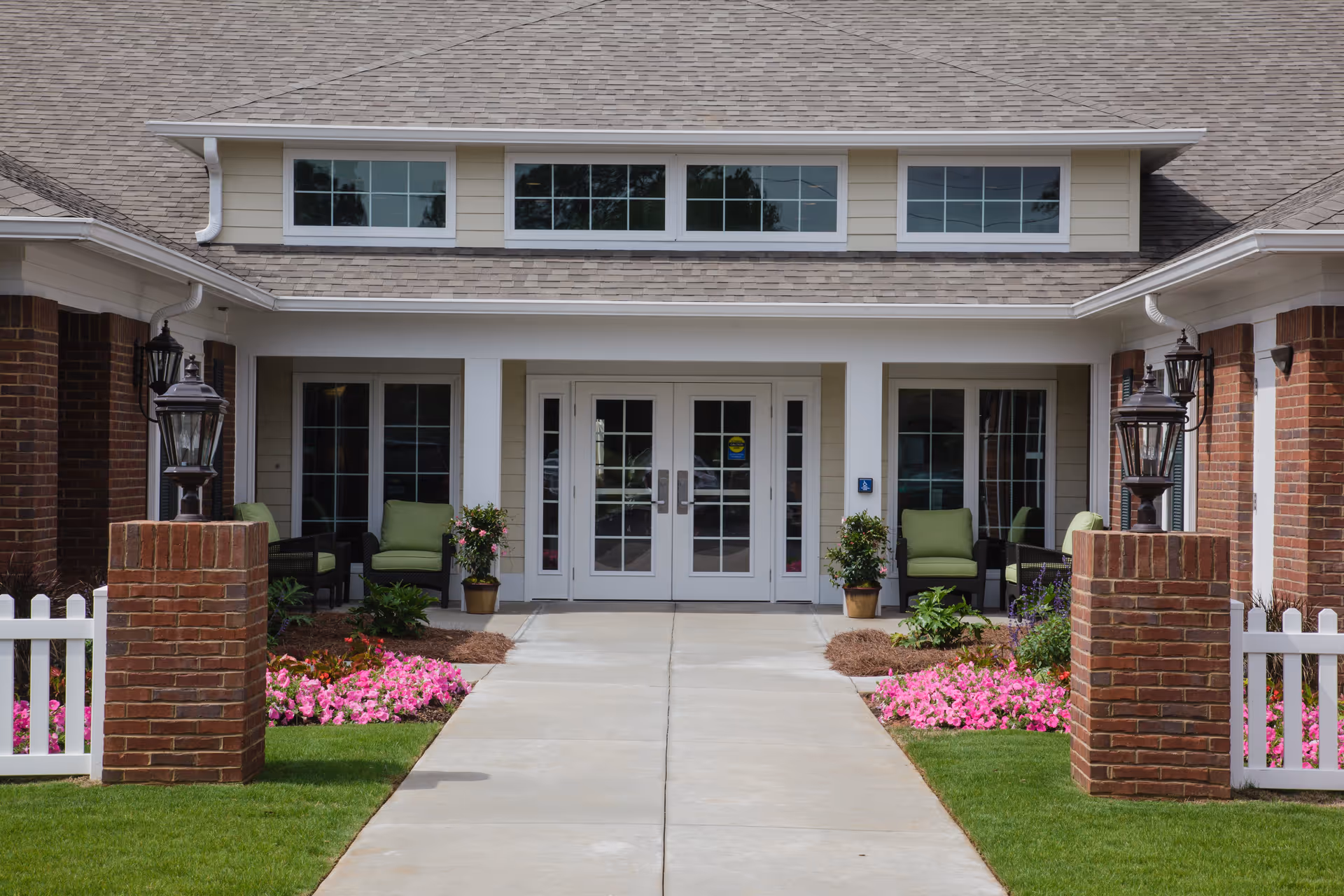 Front entrance of a senior living facility with double glass doors, flanked by windows and outdoor seating with green cushions. The entrance is framed by brick pillars with lantern-style lights, a white picket fence, and landscaped flower beds with pink flowers and green shrubs.