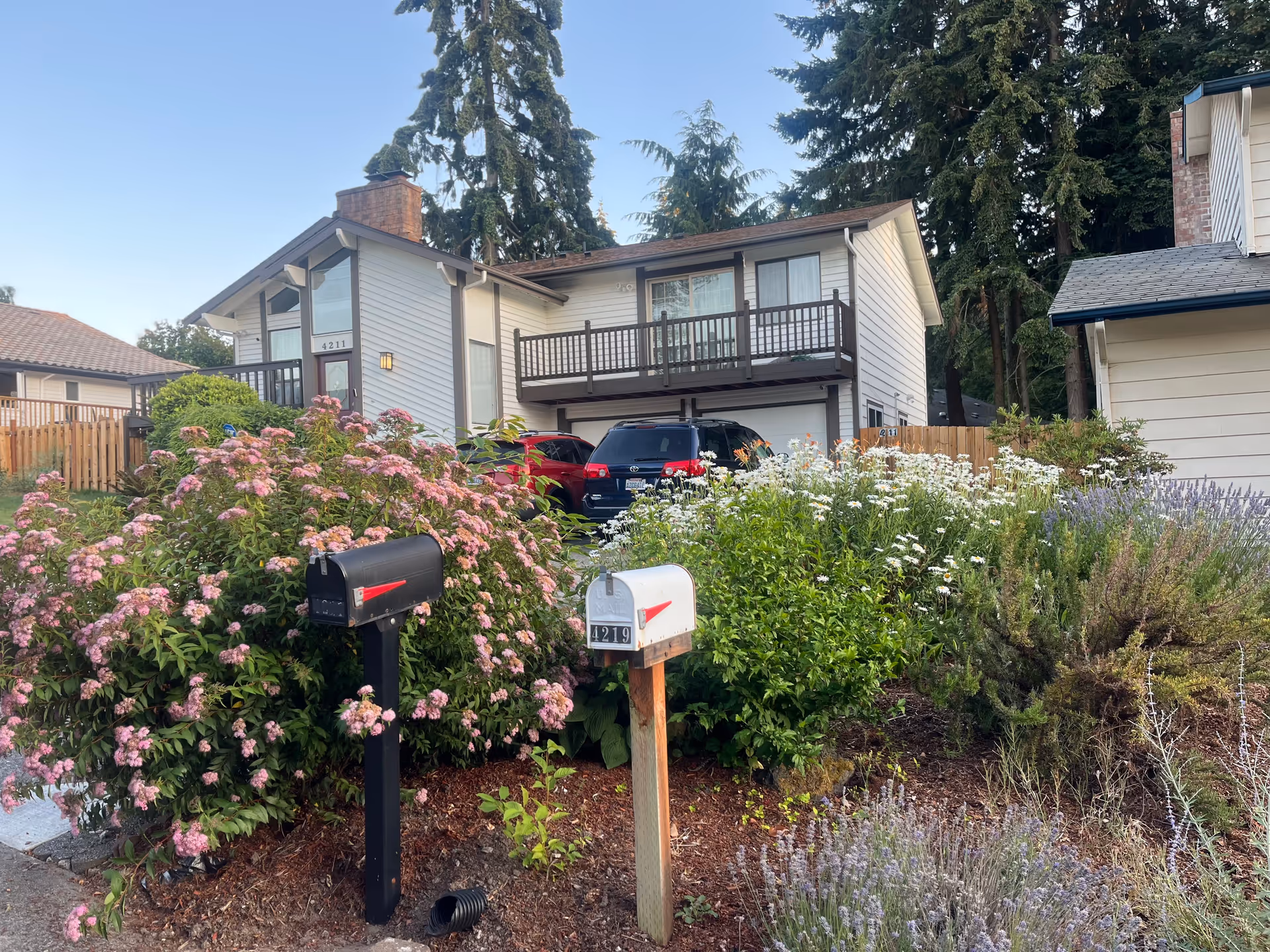 Two-story residential building with balconies, mailbox posts, and flowering garden in foreground.