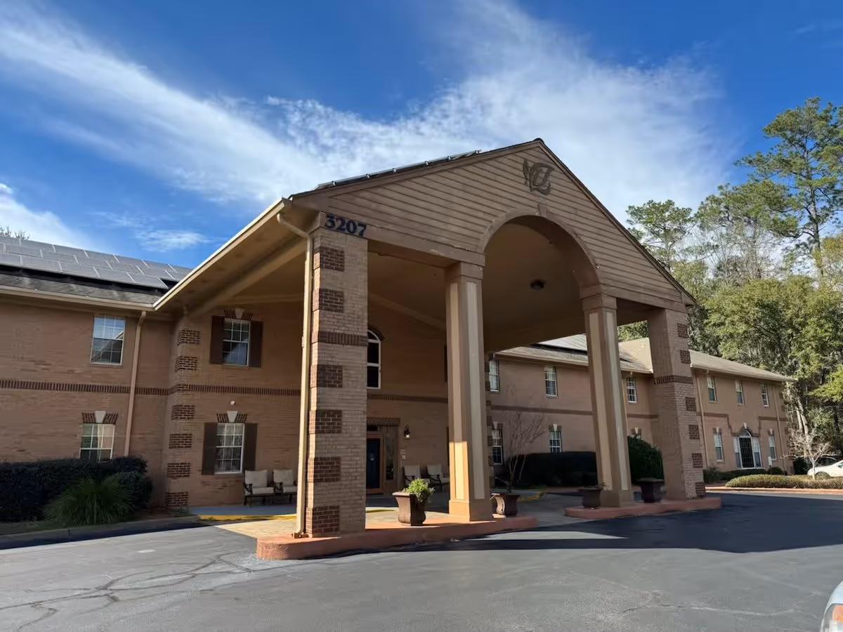 Exterior view of Woodmont Senior Living building showing a two-story brick structure with a covered entrance supported by four large columns. The building has multiple windows, some outdoor seating, potted plants near the entrance, and solar panels on the roof. Trees and a blue sky with some clouds are visible in the background.