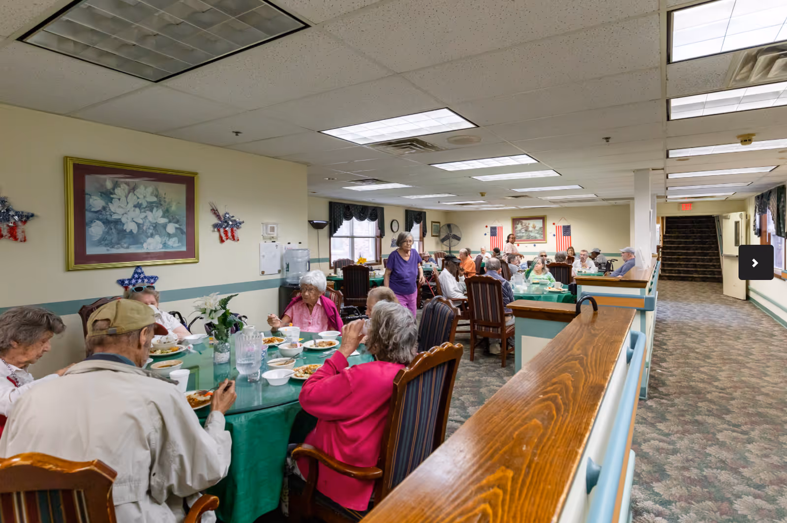 A group of elderly people sitting around tables in a dining room at Avalon Assisted Living, eating and socializing. The room has beige walls, carpeted floors, and fluorescent ceiling lights. There are patriotic decorations on the walls and a painting of flowers. A woman in a purple shirt is standing and walking among the tables.