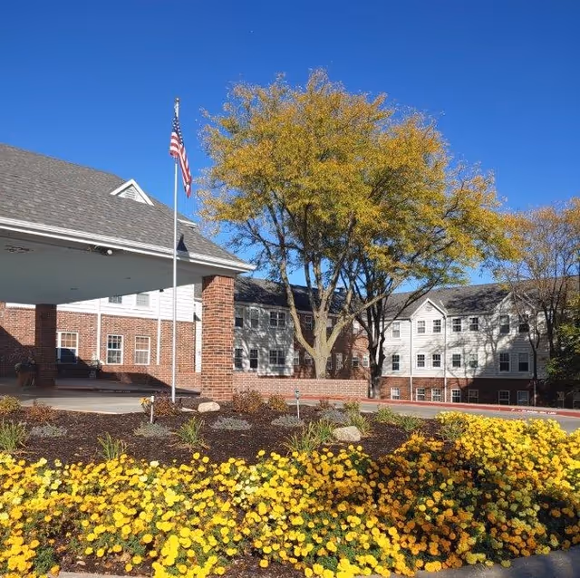 Entrance and front facade of a multi-story brick-and-white senior living building with an American flag, trees, and a bed of yellow flowers under a clear blue sky.