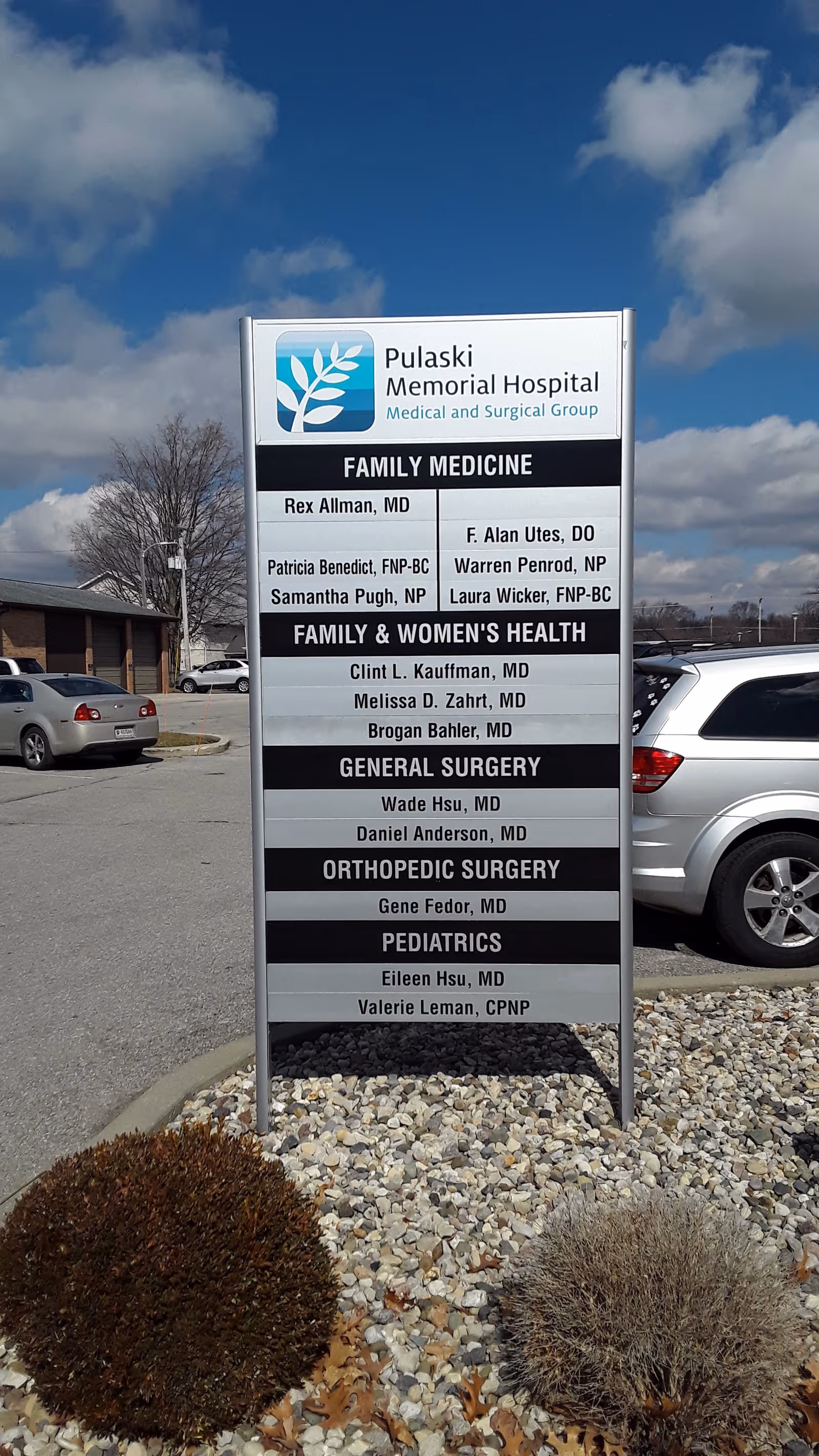 Exterior view of Pulaski Memorial Hospital sign listing departments beside parked cars.
