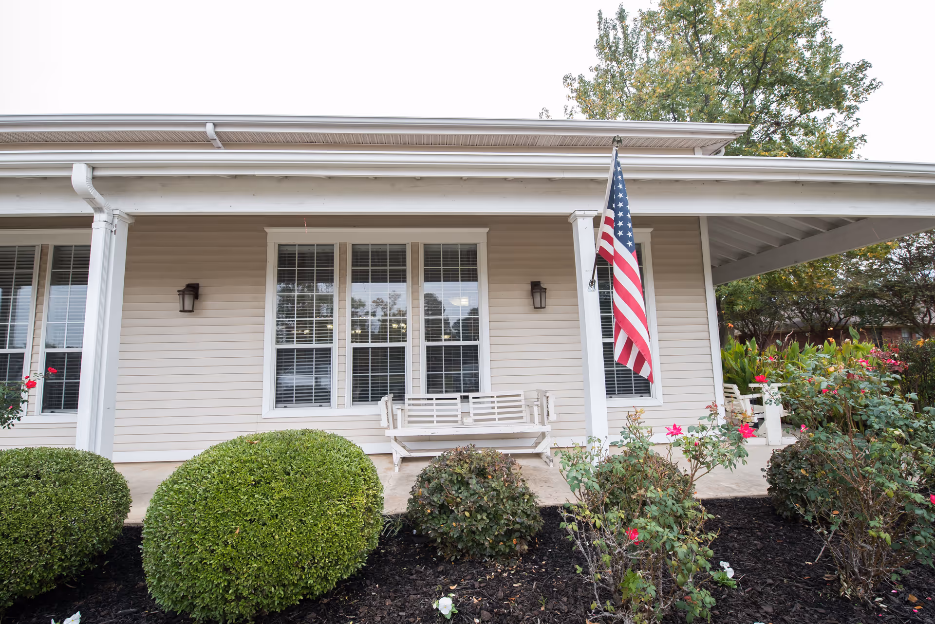 Front porch of a beige building with white trim, featuring a white wooden bench swing, an American flag hanging from a white column, two wall-mounted lantern-style lights, and neatly trimmed bushes and flowering plants in the garden bed in front.