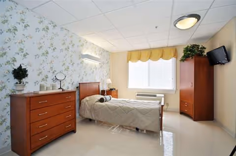 Senior living bedroom with bed, dresser, wardrobe, window, and floral wallpaper.