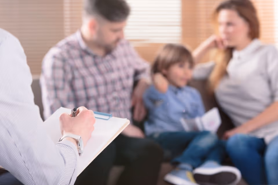 Staff member fills out a clipboard while a family discusses care in a living room.