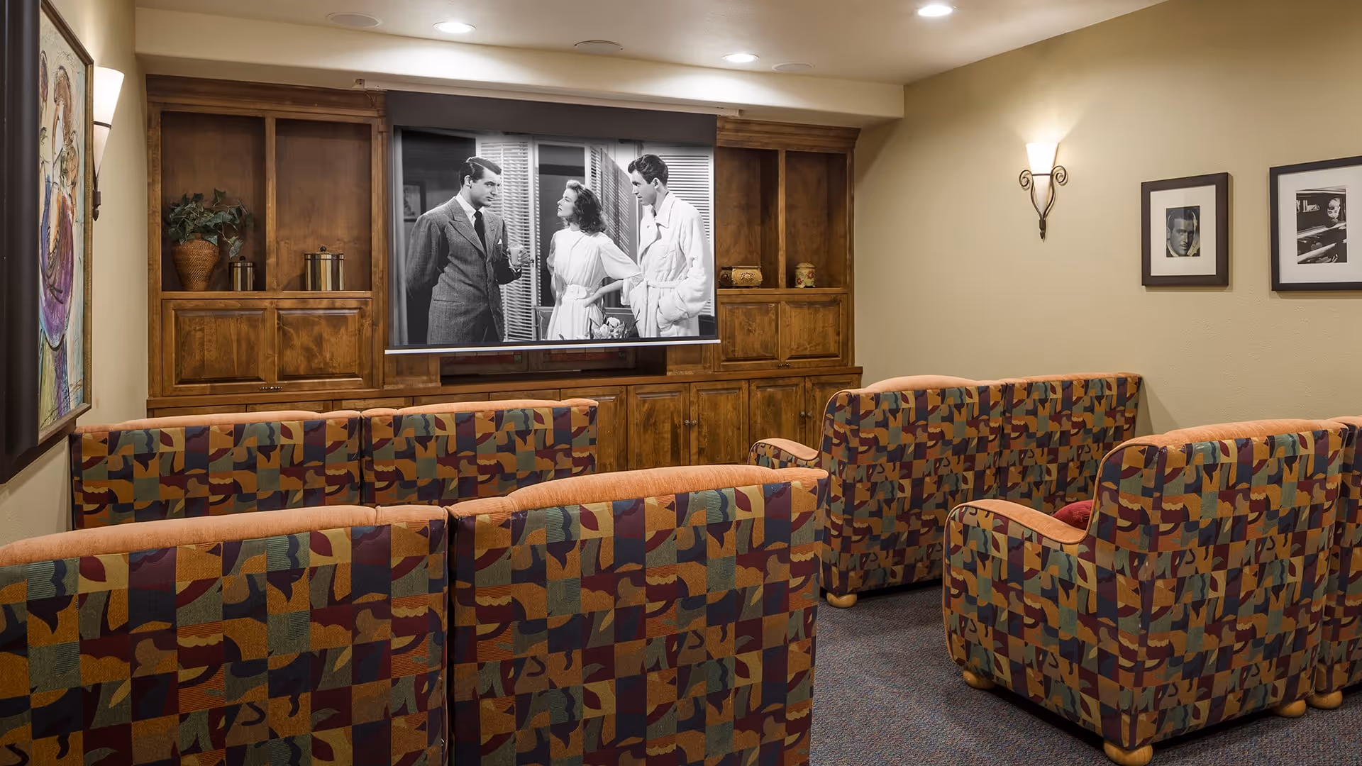 A cozy media room with patterned upholstered chairs arranged in rows facing a large screen displaying a black-and-white movie scene. The room has wooden built-in cabinets, framed black-and-white photos on the wall, and warm lighting.