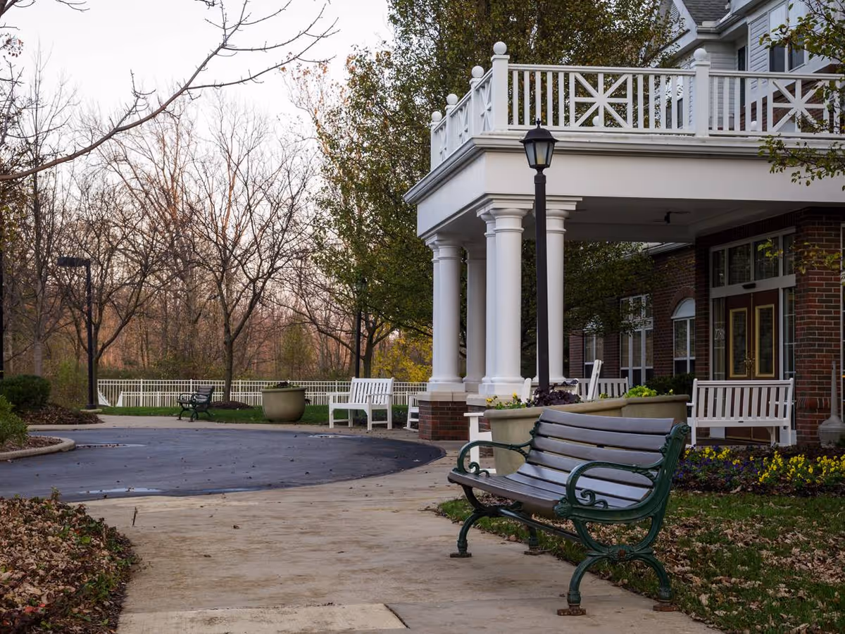 Front entrance of a senior living building with a covered porch supported by white columns, benches, and surrounding trees.