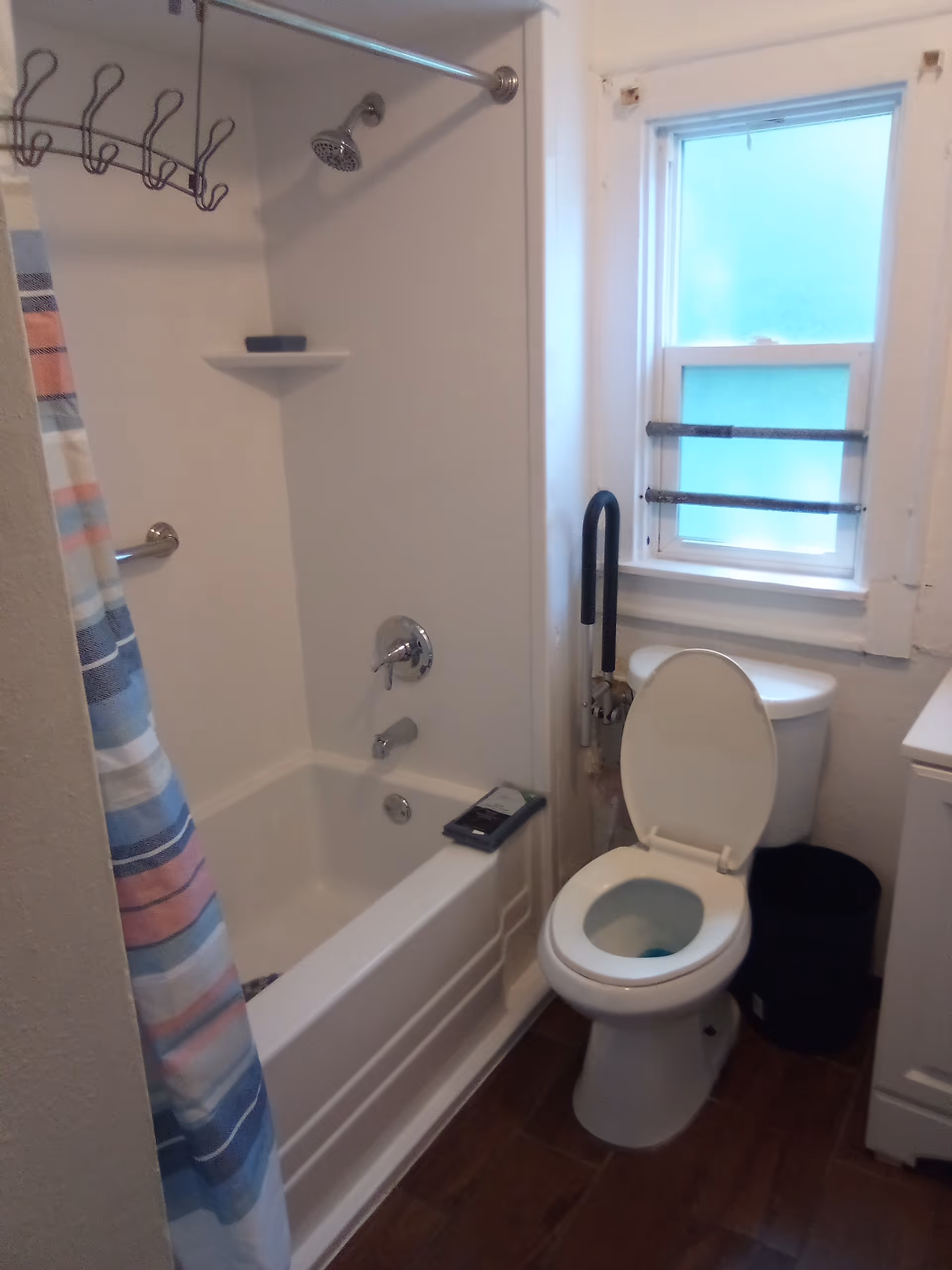 Small bathroom with shower/tub, striped curtain, toilet, window, and safety rail.