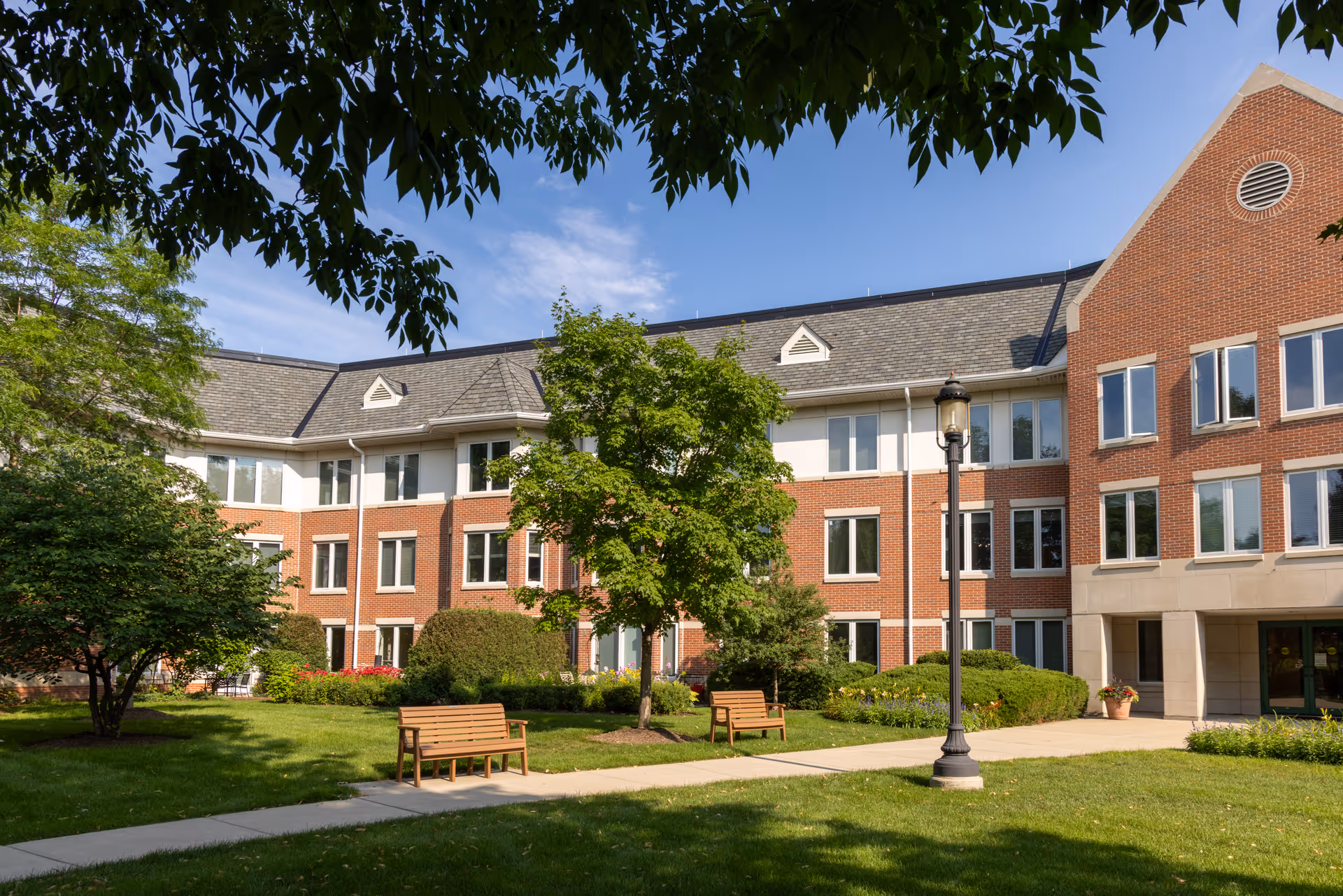 Brick senior living building front with a landscaped courtyard, benches, trees, and a lamp post.