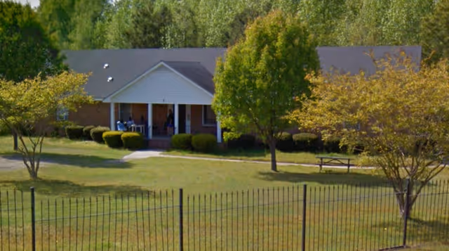 Single-story brick assisted living building with a covered front porch, lawn, trees, and a black metal fence in the foreground.