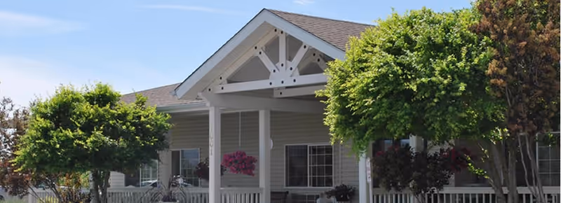 Front exterior view of a senior living facility building with a covered entrance, white pillars, and a gabled roof. There are green trees and shrubs in front of the building.