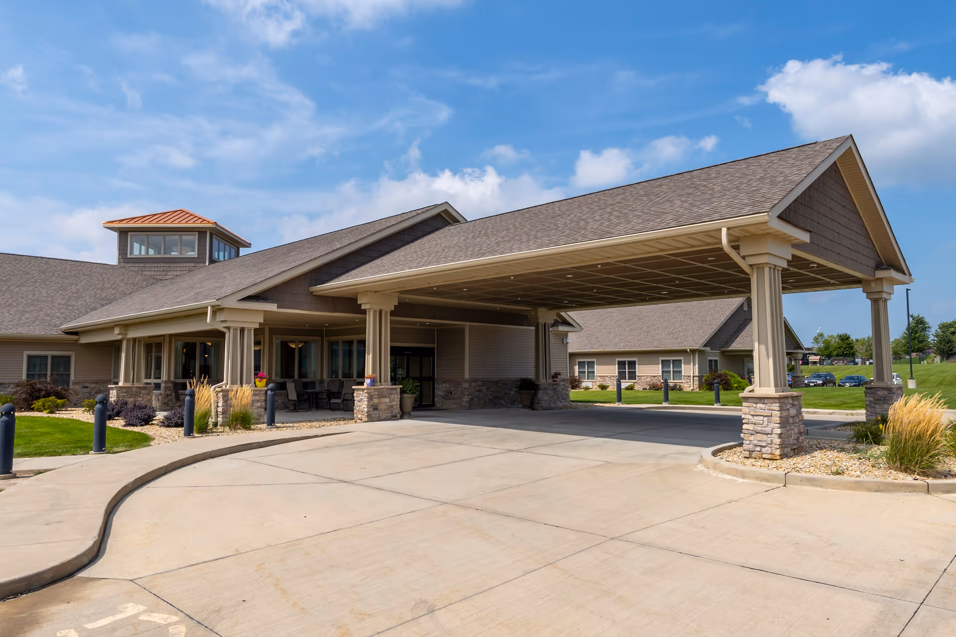 Exterior view of The Carriages of Bloomington senior living facility showing the main entrance with a covered drop-off area supported by columns with stone bases, surrounded by landscaped greenery and a clear blue sky.