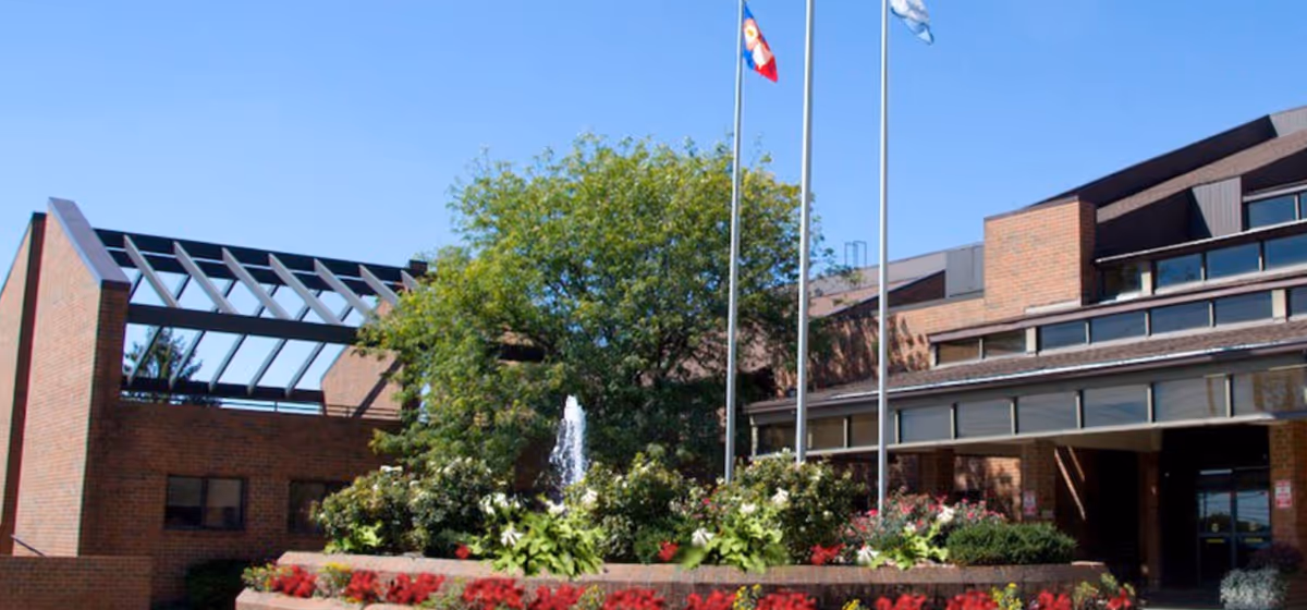 Front entrance of a brick retirement community building with flagpoles, a fountain, and landscaped flower beds.