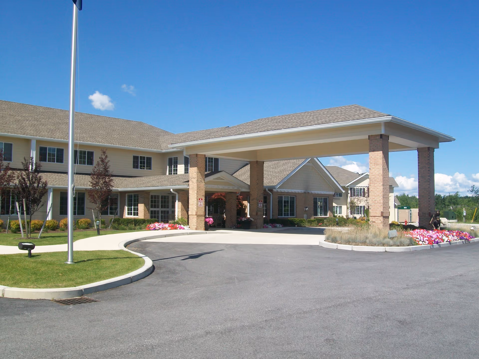 Exterior view of Winterberry Heights Senior Living facility showing a large covered entrance with brick pillars, a driveway, landscaped flower beds, and a clear blue sky.