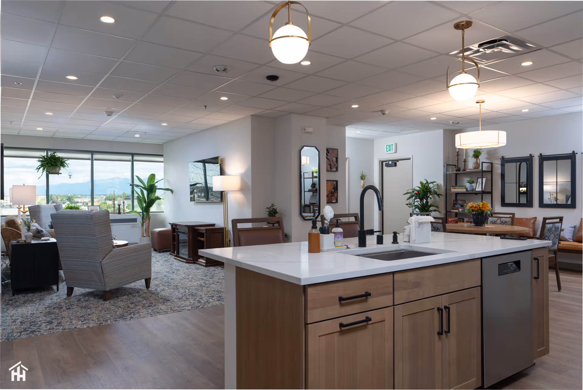 A spacious and modern common area in a senior living facility featuring a kitchen island with a sink and faucet in the foreground. The room includes comfortable seating with armchairs and sofas, large windows with a scenic view, decorative plants, and warm lighting fixtures hanging from the ceiling.
