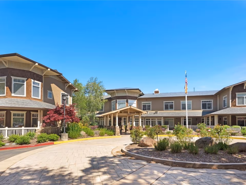 Exterior view of Sunrise of Fair Oaks senior living facility showing a two-story building with beige and brown siding, multiple windows, a covered entrance, landscaped garden with shrubs and rocks, and a flagpole with an American flag under a clear blue sky.