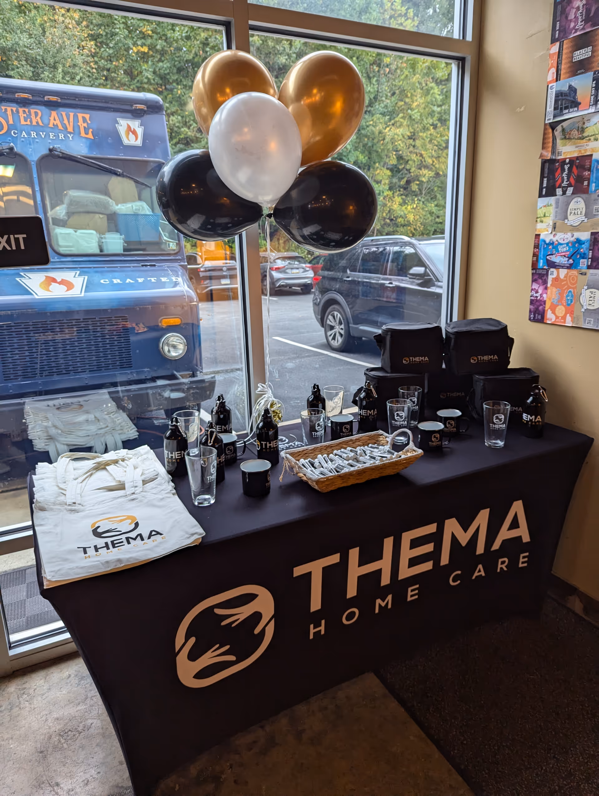 Promotional table with Thema Home Care branding, balloons, and freebies inside a facility lobby.