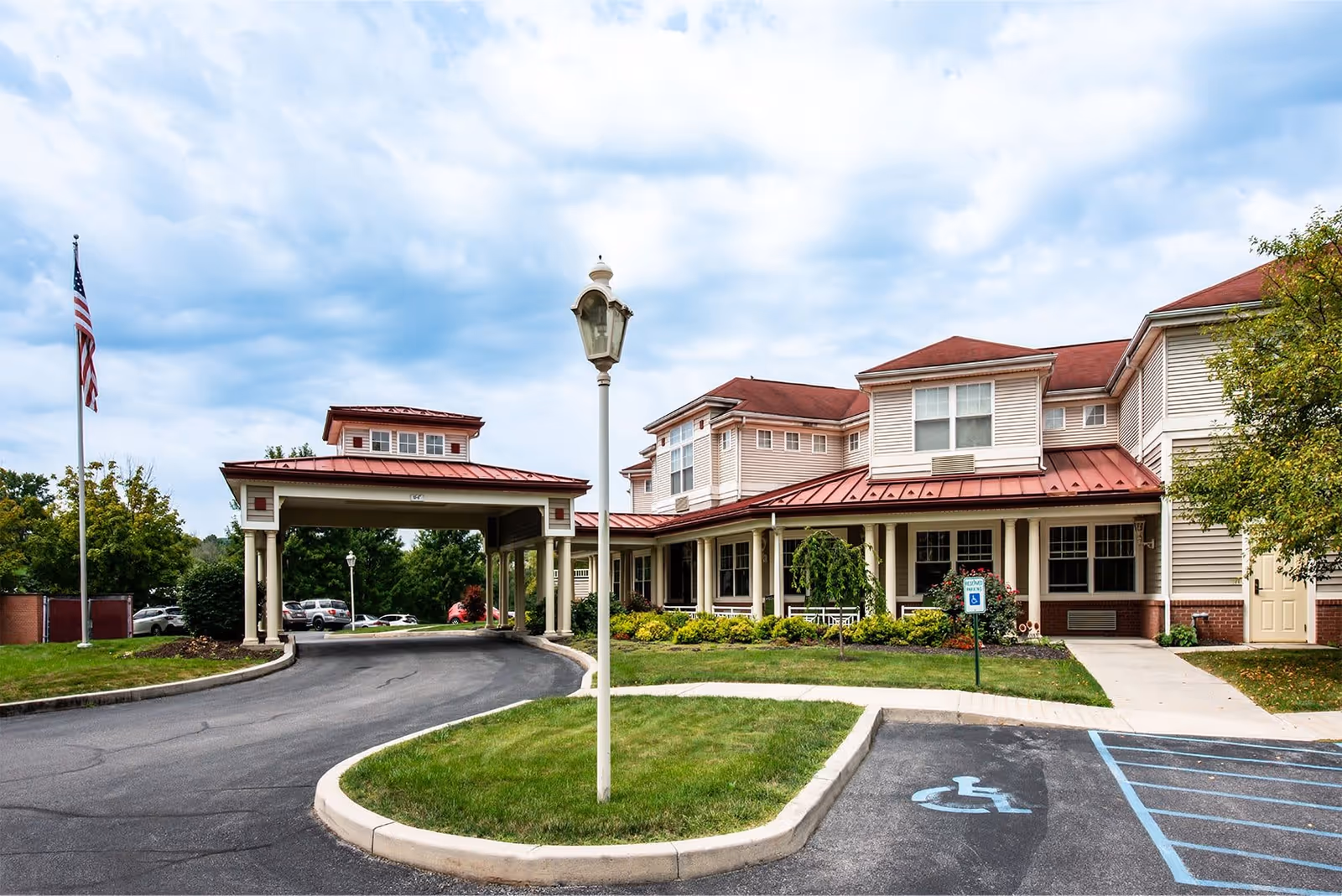 Exterior view of The Winds At Mattern Orchard by New Perspective, showing a two-story building with beige siding and red roofs. There is a covered driveway entrance, a flagpole with an American flag, a vintage-style street lamp, landscaped greenery, and a handicapped parking space in the foreground.