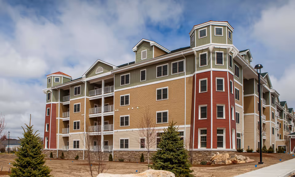 Four-story multi-colored senior living building with balconies and turret-like towers under a partly cloudy sky.