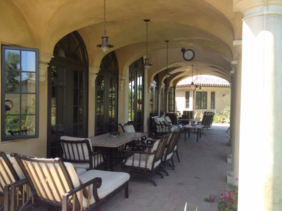A covered outdoor patio with arched ceilings, columns, and multiple cushioned dining and lounge chairs arranged around tables.