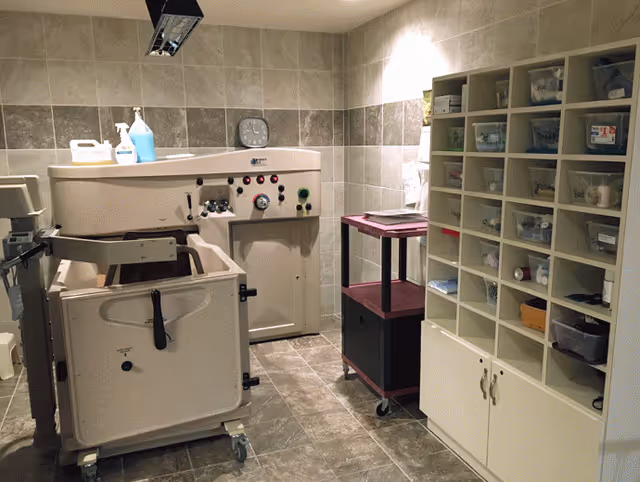 Assisted bathing room with a large mechanical tub, tiled walls and floor, storage cubbies filled with supplies, and a supply cart.