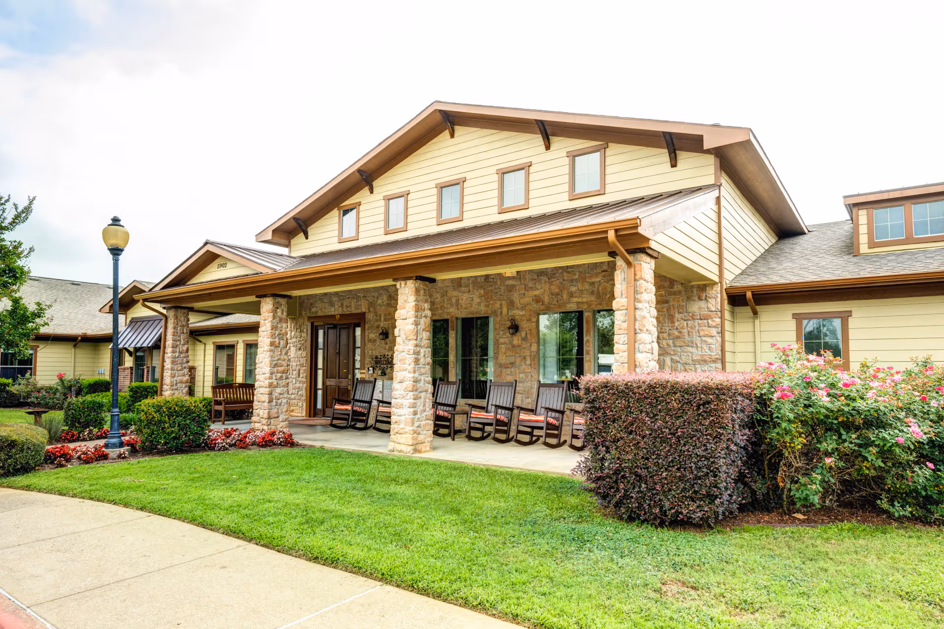Front exterior view of a single-story assisted living facility with stone pillars supporting a covered porch. The porch has several wooden rocking chairs with cushions. The building has beige siding with brown trim and multiple windows. There is a well-maintained lawn, bushes, and flowers in front of the building, along with a sidewalk and a streetlamp.
