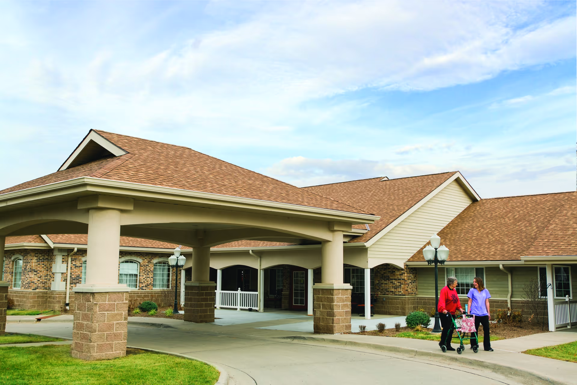 Exterior view of a senior living facility entrance with a covered drop-off and two pedestrians.
