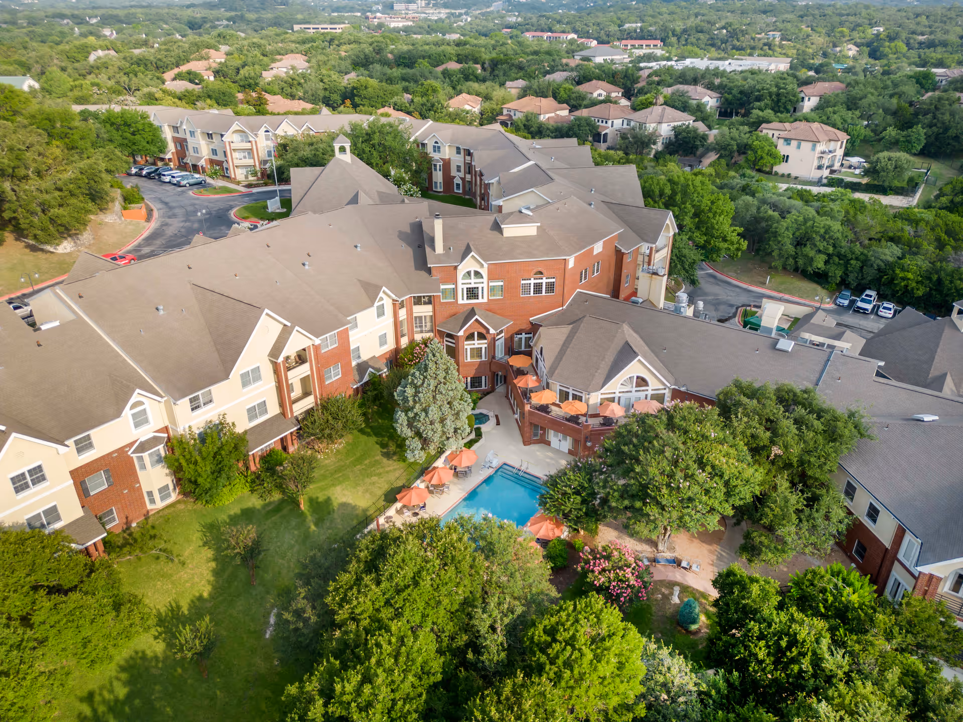 Aerial view of Brookdale Westlake Hills senior living facility showing multiple connected buildings with beige and red brick exteriors, surrounded by green trees and landscaping. The facility features an outdoor swimming pool with orange umbrellas and lounge chairs. Parking areas and neighboring residential buildings are visible in the background.