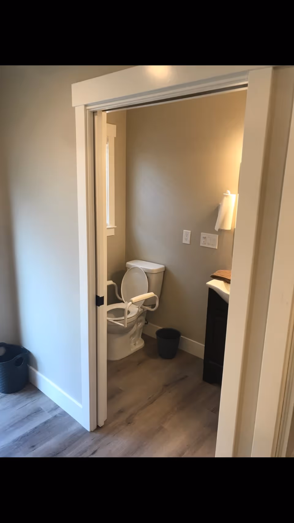 Small accessible bathroom showing a toilet with white safety rails, a sink vanity, and a waste bin.