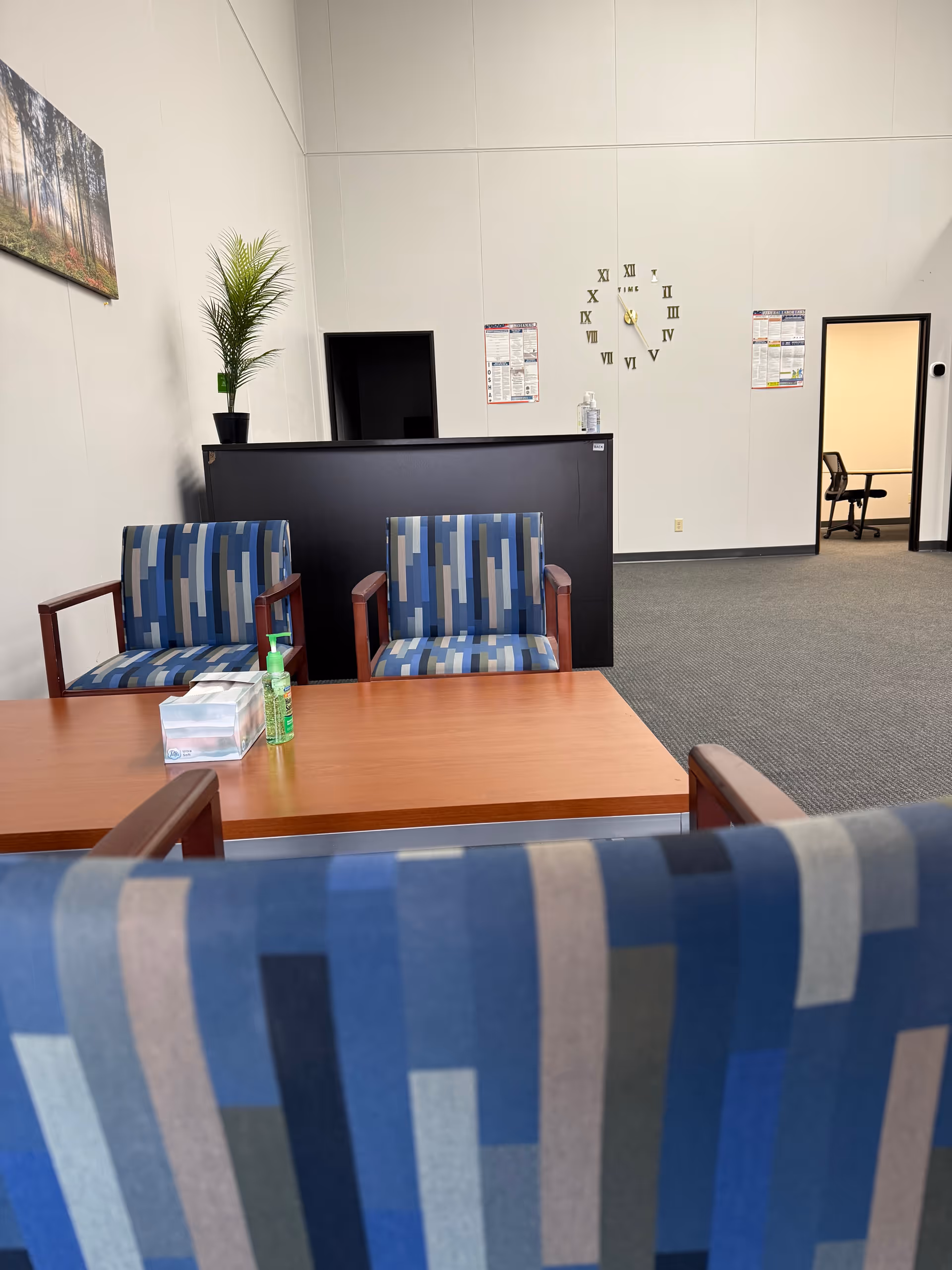 A reception area with blue patterned chairs, a wooden table, and a wall clock.