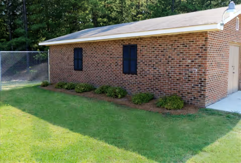 Single-story brick building exterior with green lawn, shrubs, and a chain-link fence.