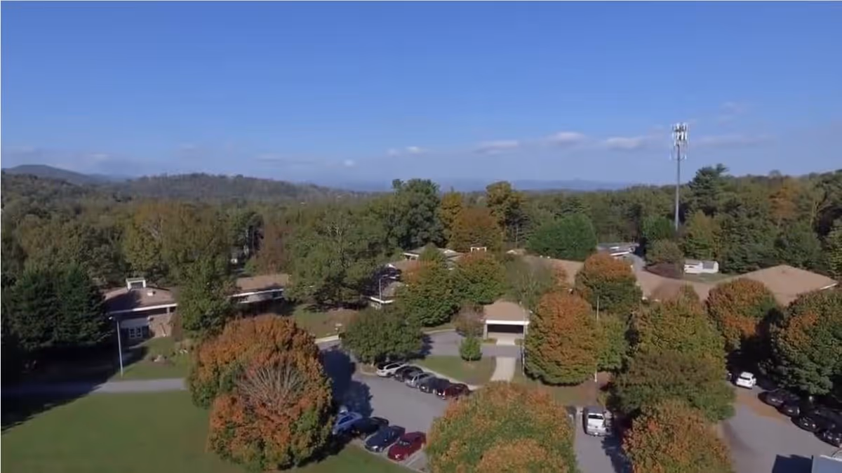 Aerial view of River Bend Assisted Living facility surrounded by trees with autumn foliage. Several buildings with brown roofs are visible along with parked cars in the parking lot. The background shows a forested area and distant hills under a clear blue sky.