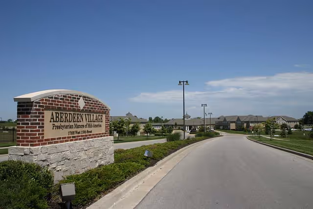 Entrance sign reading 'Aberdeen Village' beside a curved driveway leading to the senior living campus under a clear blue sky.