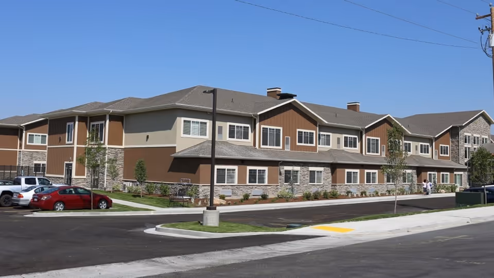 Two-story senior living building with brown beige siding and a surrounding parking lot.