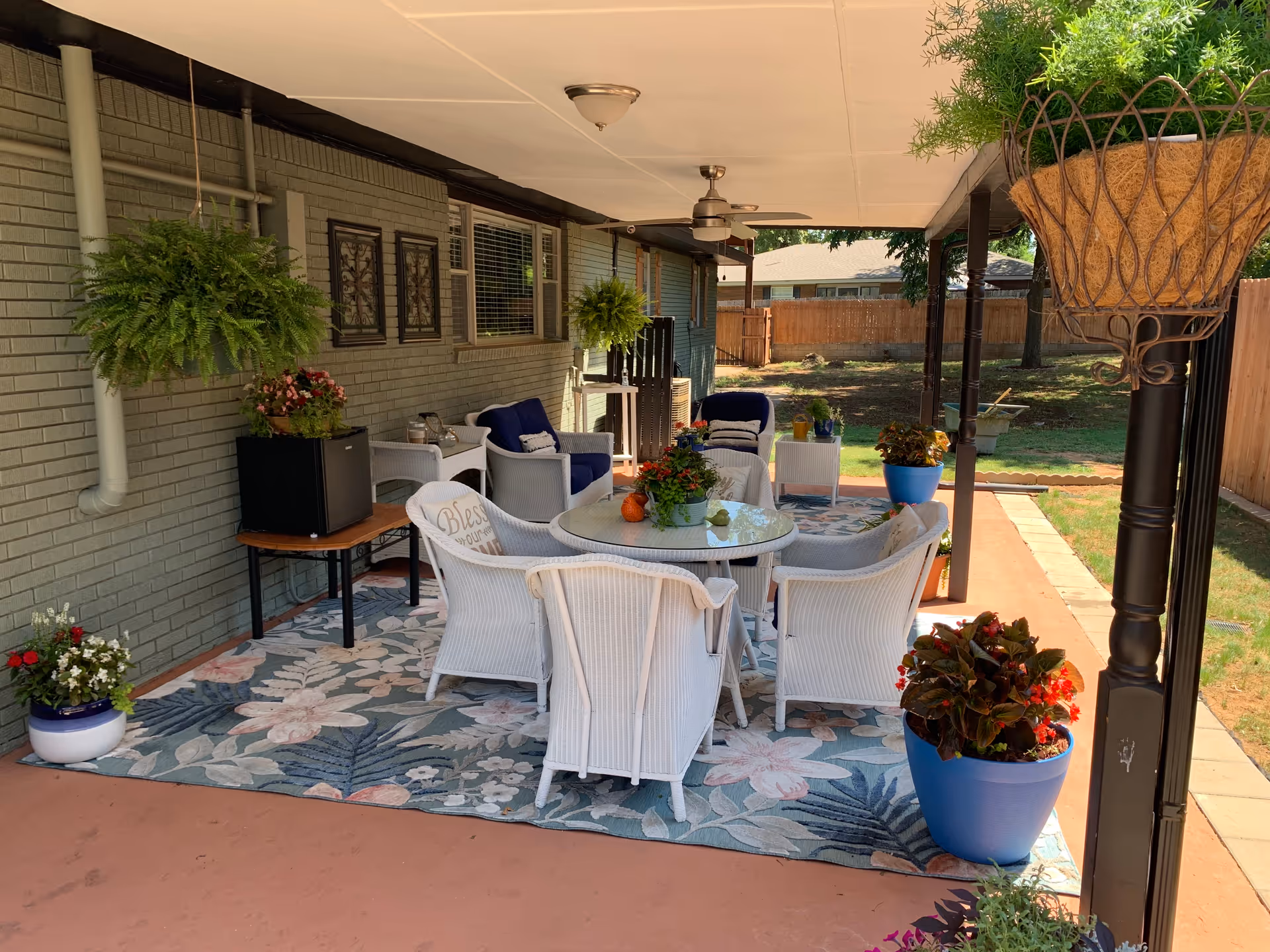 Outdoor covered patio with white wicker chairs around a glass table and potted plants.