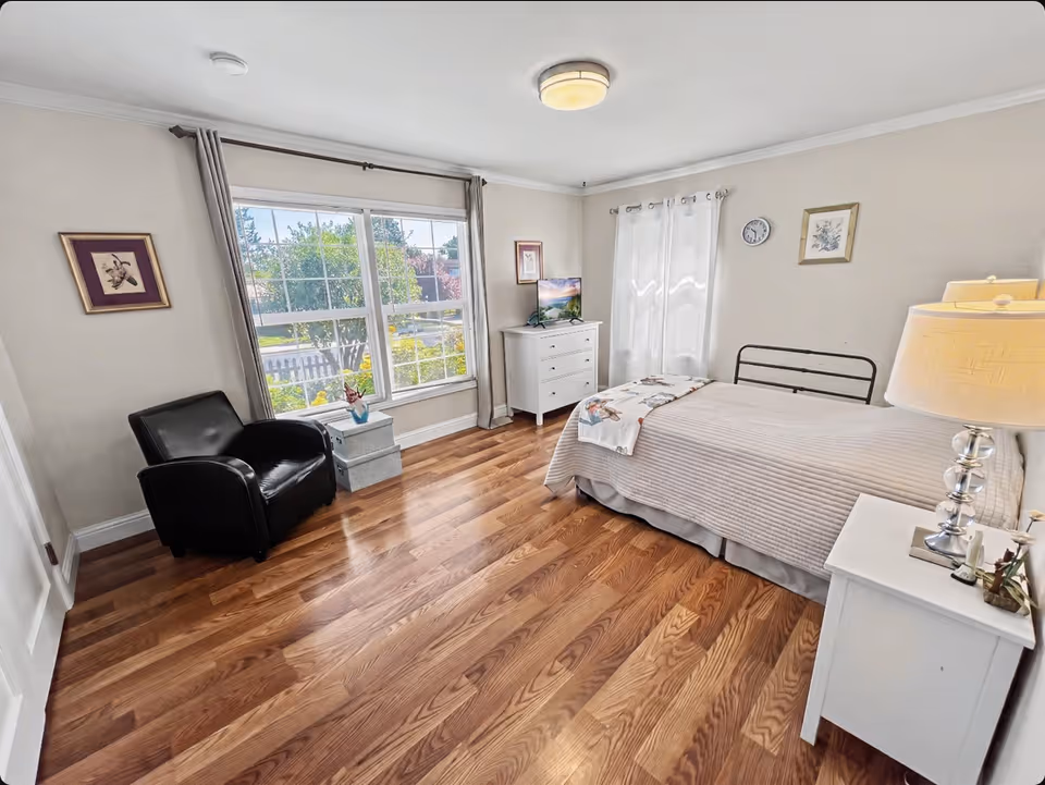 Bright, neatly arranged bedroom with a bed, dresser, chair, and large sunlit window.