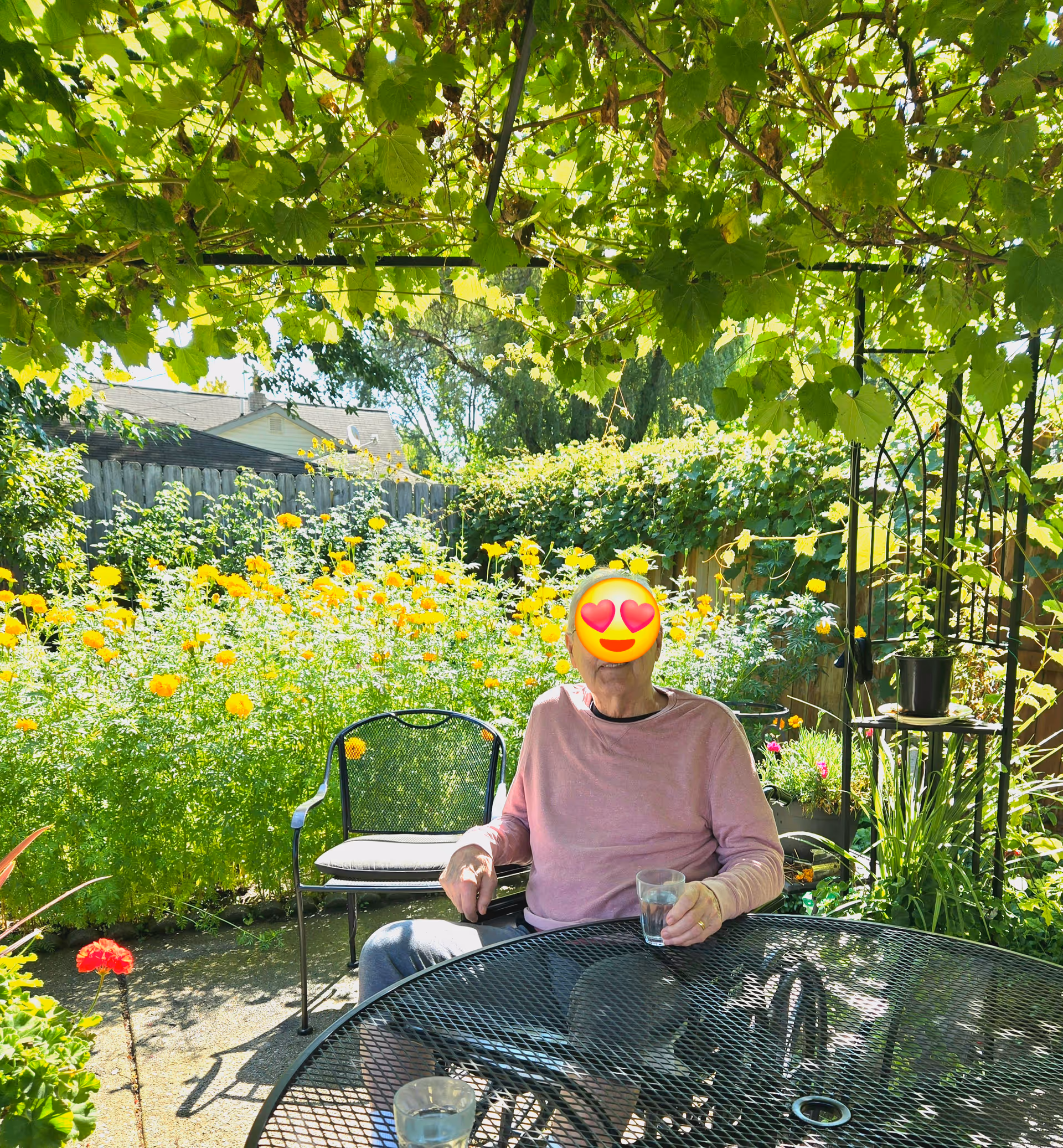 A resident sits at a metal patio table in a lush garden with yellow flowers.