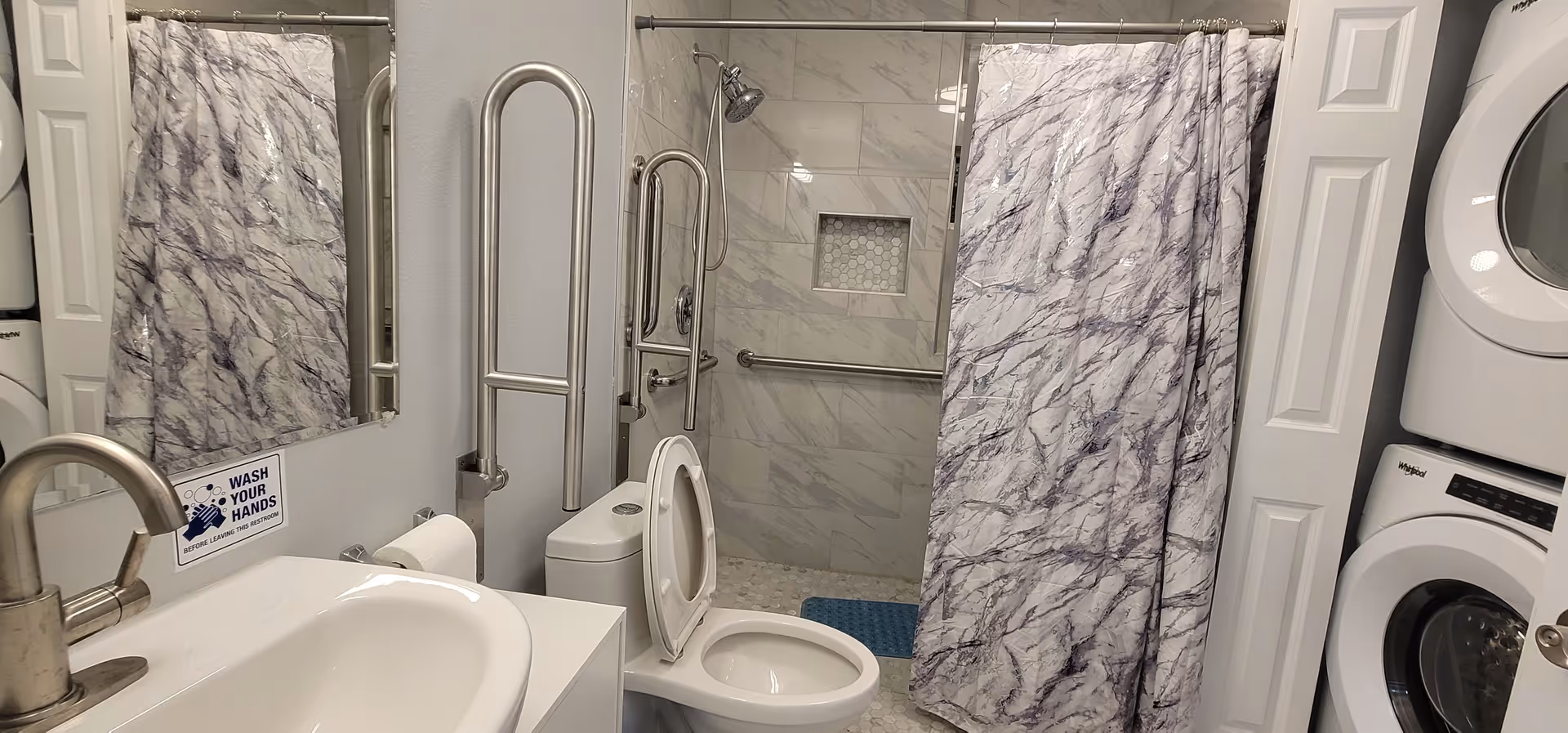 Accessible bathroom with toilet, sink, shower, grab bars, marble walls, and stacked washer and dryer.