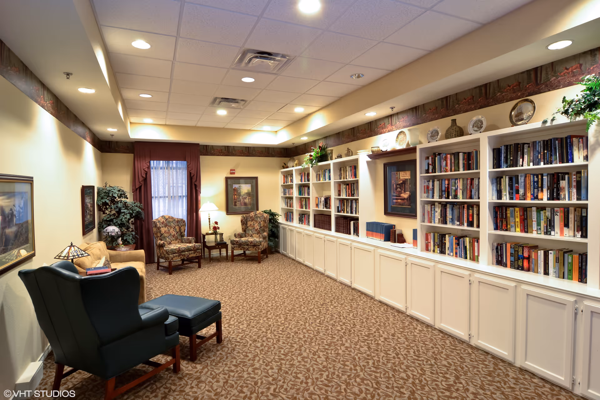 A cozy senior living facility library or reading room with built-in white bookshelves filled with books, floral patterned armchairs, a blue armchair with matching ottoman, a beige sofa, framed artwork on the walls, potted plants, and warm lighting.