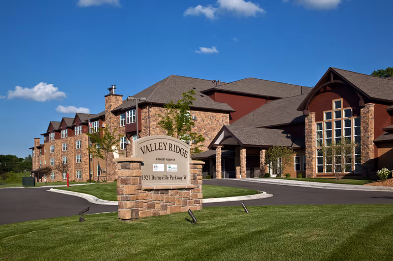 Stone entrance sign reading 'Valley Ridge' sits on a lawn in front of a large brick-and-wood senior living building under a blue sky.
