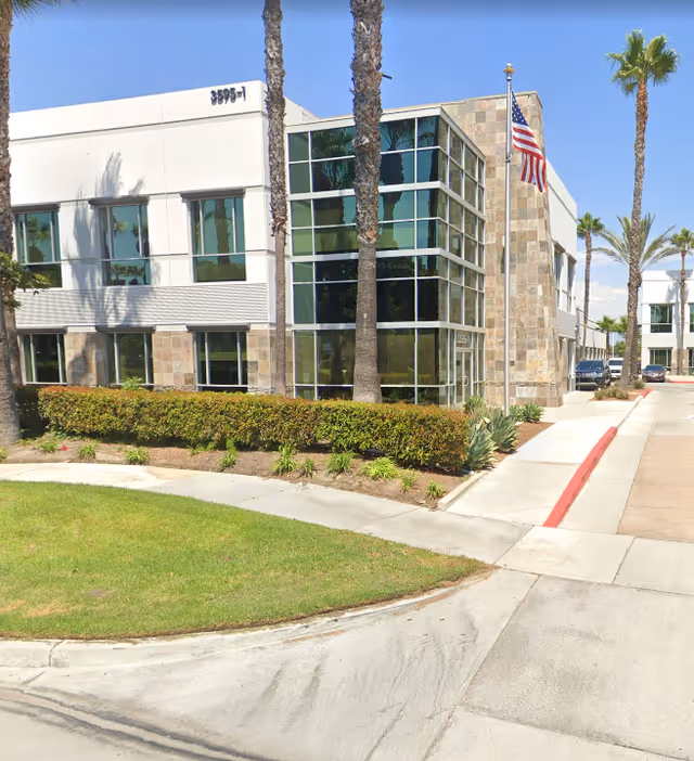 Modern senior living facility exterior with glass windows, palm trees, and an American flag.