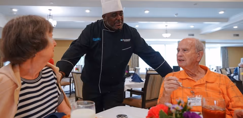A chef in a black uniform and white hat is talking to two elderly residents seated at a dining table in a well-lit dining room. The woman on the left is wearing a striped shirt and beige jacket, while the man on the right is wearing an orange shirt and holding a spoon. The table has drinks and a small floral centerpiece.